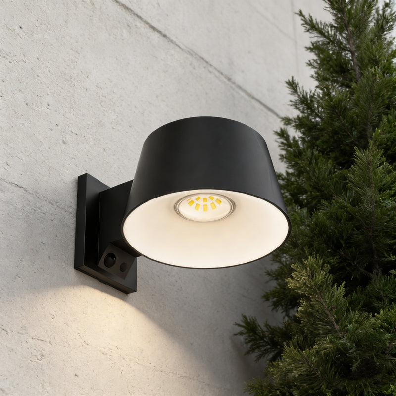 A modern black outdoor wall sconce with a cylindrical shade, mounted on a textured concrete wall next to a green evergreen tree. The light is turned on, casting a warm glow downward.