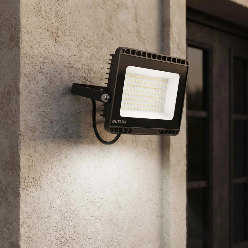 A black OUTLUX LED wall-mounted floodlight attached to a textured beige stucco wall, casting light downward, with a dark doorway in the background.