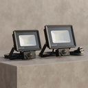 Two OUTLUX outdoor floodlights in matte black, shown side-by-side on a textured gray concrete block with their power cords and plugs visible.