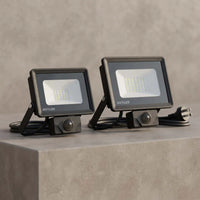 Two OUTLUX outdoor floodlights in matte black, shown side-by-side on a textured gray concrete block with their power cords and plugs visible.