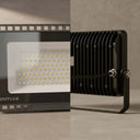 A black OUTLUX LED floodlight shown in a split close-up: the left half shows the front face with many small LED chips and a white label area, and the right half shows the side with ribbed metal heat dissipation, a mounting bracket, and a hose connector. The product is photographed on a neutral gray surface and backdrop.